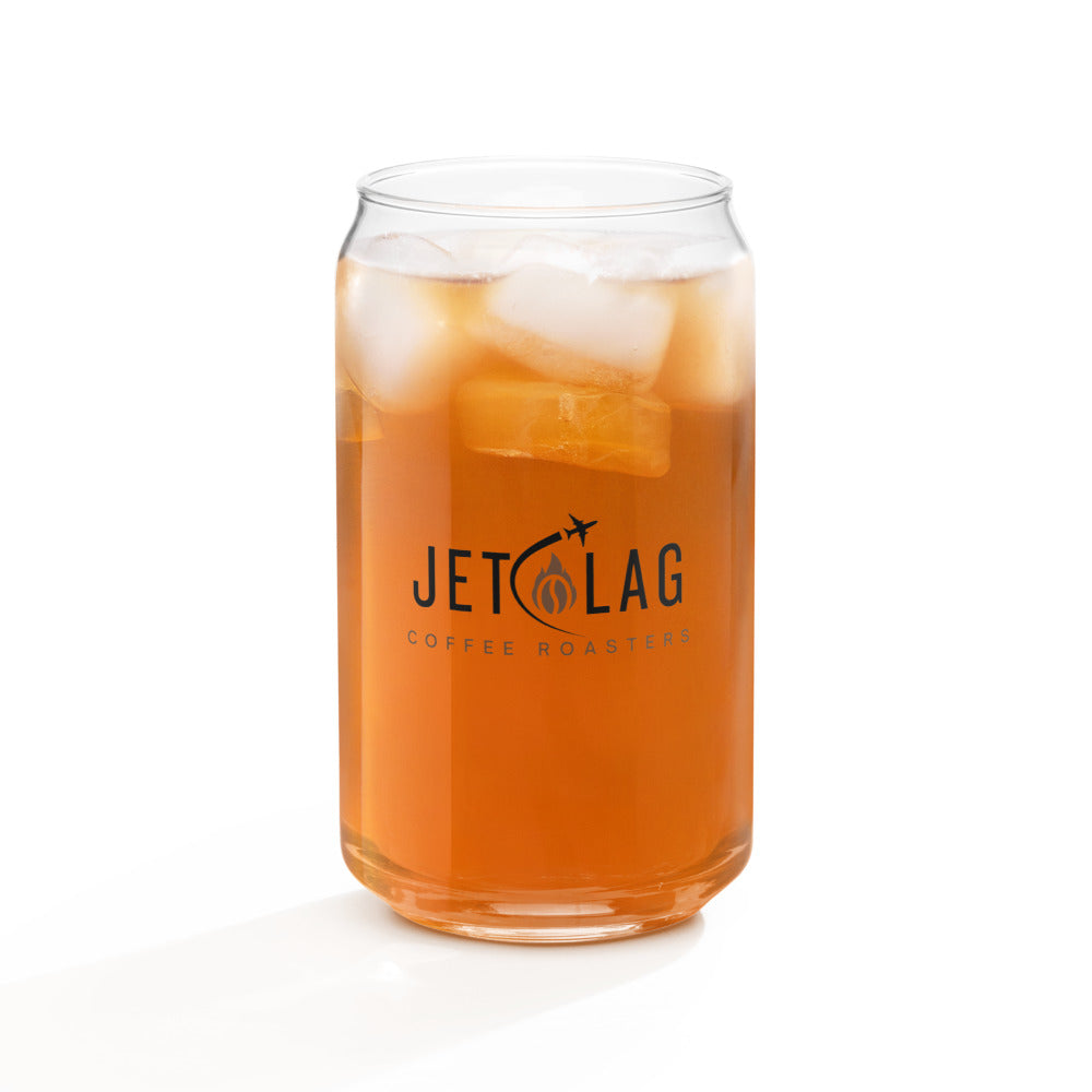 The image shows a clear glass shaped like a soda can filled with an iced beverage. The drink appears to be a cold tea or coffee, with visible ice cubes inside. The glass features the logo for "Jet Lag Coffee Roasters," which includes the text "JET LAG" with an illustration of two small airplanes and a stylized coffee bean and flame. The words "COFFEE ROASTERS" are printed below the main logo. The glass is set against a white background.